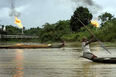 photo of a woman in a small boat in the foreground, paddling; in the background jets of flames from refinery stacks are nearby