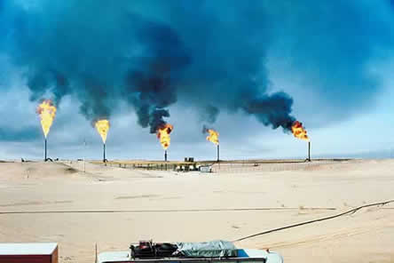 photo of a refinery with flames coming from towers in a barren landscape. the sky is darkened by the smoke.